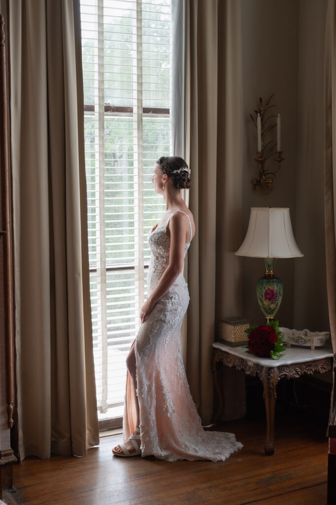 Bride in a lace wedding gown standing by tall windows with sheer blinds, natural light on her profile.