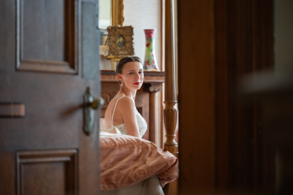 Bride in a white lace wedding dress sits on a bed in a warmly lit bedroom, gazing toward the camera from a doorway.