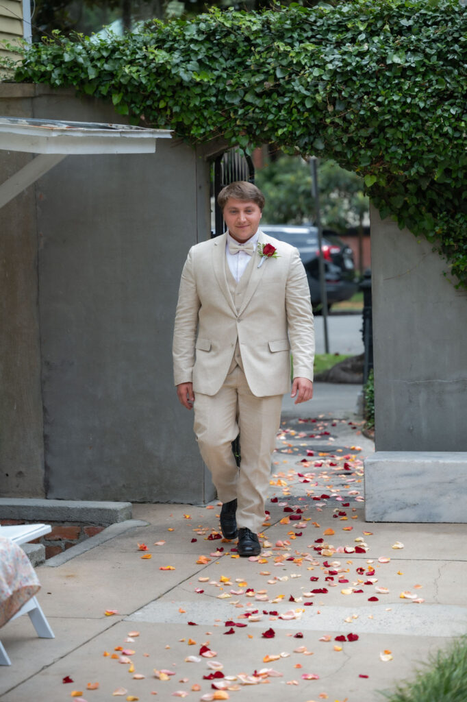 Man in a light beige suit walking down a sidewalk, boutonniere on lapel, rose petals scattered on ground under a green leafy archway.
