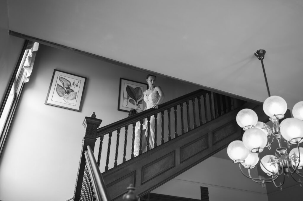 Bride on a grand staircase landing in a lace wedding gown, chandelier visible in the foreground and botanical art on the wall.