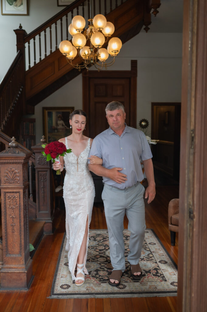 Bride in a white lace wedding dress holds a red bouquet while an older man escorts her down a wood-paneled hallway under a brass chandelier in an ornate home interior.
