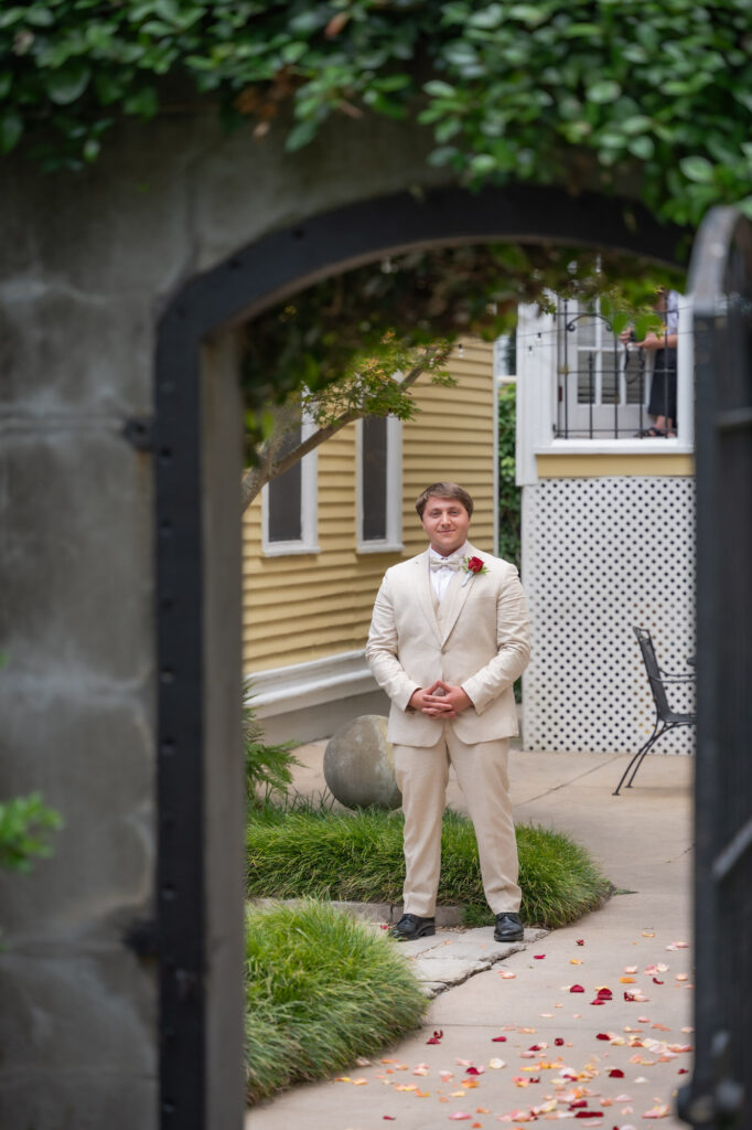 Man in a beige suit with a boutonniere stands outdoors under an arch, petals scattered on the walkway across the path.