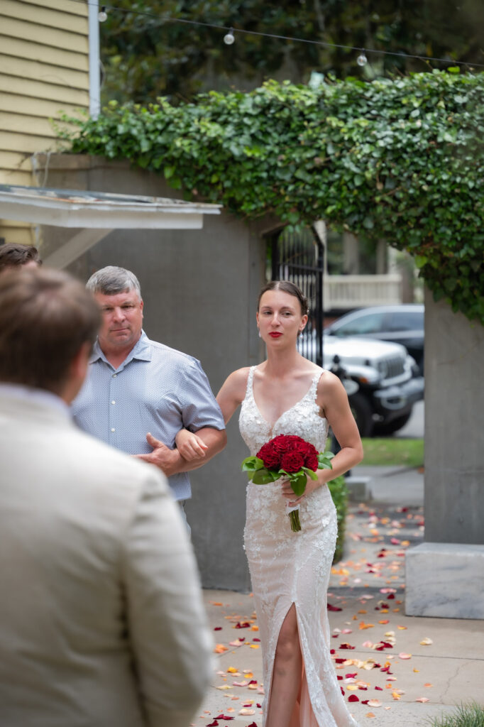 Bride in a white lace wedding dress holding a red bouquet, walking arm‑in‑arm with a man along a petal‑strewn sidewalk.