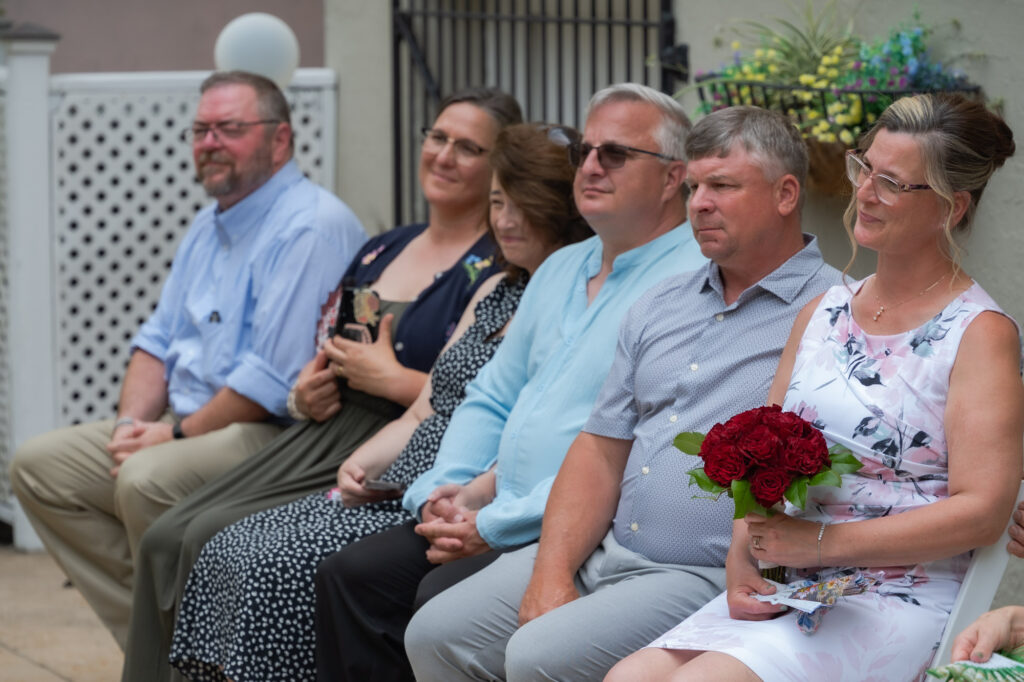 Seven adults seated outdoors in a line; the woman on the far right wears glasses and holds a red bouquet.