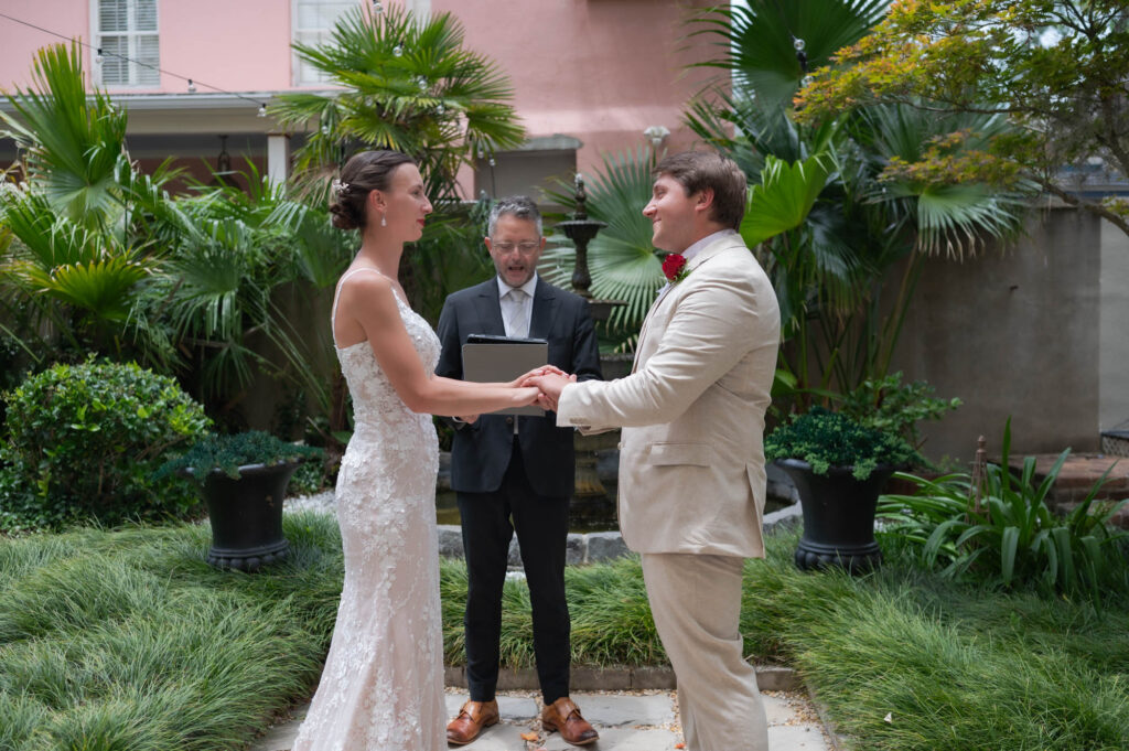 Bride in a lace gown and groom in a light suit hold hands, facing each other as an officiant reads vows in a lush garden setting.