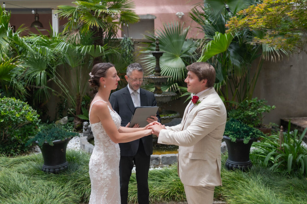 Bride and groom hold hands while an officiant reads vows in a lush garden setting with a fountain behind them.