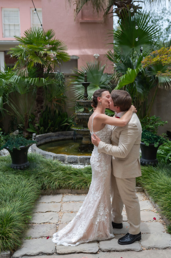 Bride in a lace wedding gown and groom in a beige suit kiss by a stone fountain in a tropical garden setting.