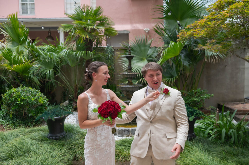 Bride in a white lace dress holds a red bouquet as she dances with a man in a light tan suit in a lush garden setting with a fountain in the background.