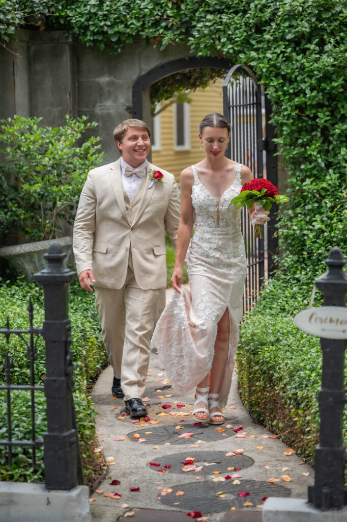 Bride in lace white wedding dress holds red bouquet as she and a groom in a beige suit walk along a petal-strewn garden path under a hedged archway.