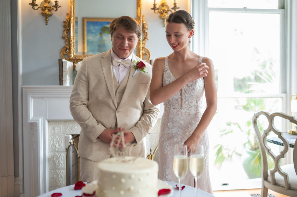 Bride in a lace dress and groom in a beige suit stand by a white wedding cake, smiling before cutting it.