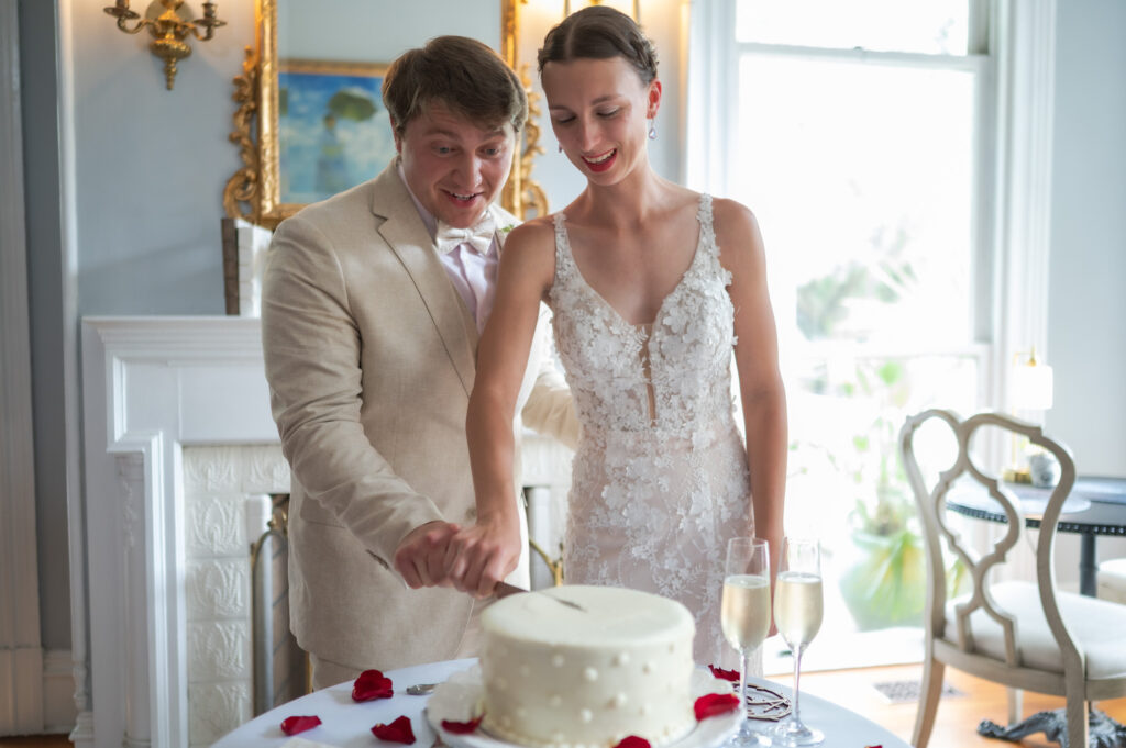 Bride and groom cut a white wedding cake together, with champagne flutes on the table nearby in a bright room.Transition of celebration is visible with petals on the cake plate.