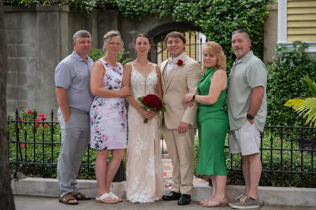Bride and groom pose with family outdoors; bride in lace gown holding red bouquet, groom in beige suit with red rose boutonniere, surrounded by relatives in pastel and green outfits.