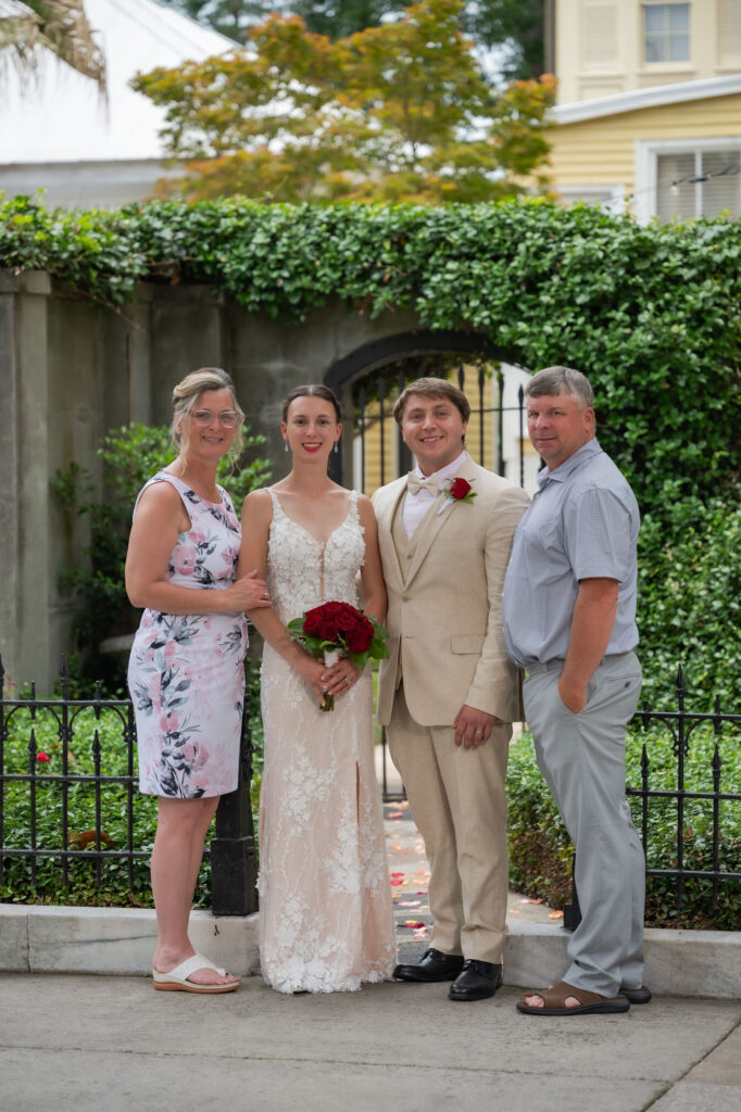 Bride in a lace white gown holding a red bouquet, flanked by her family outdoors by a ivy-covered wall and arched gate, with two men and a woman in a floral dress.