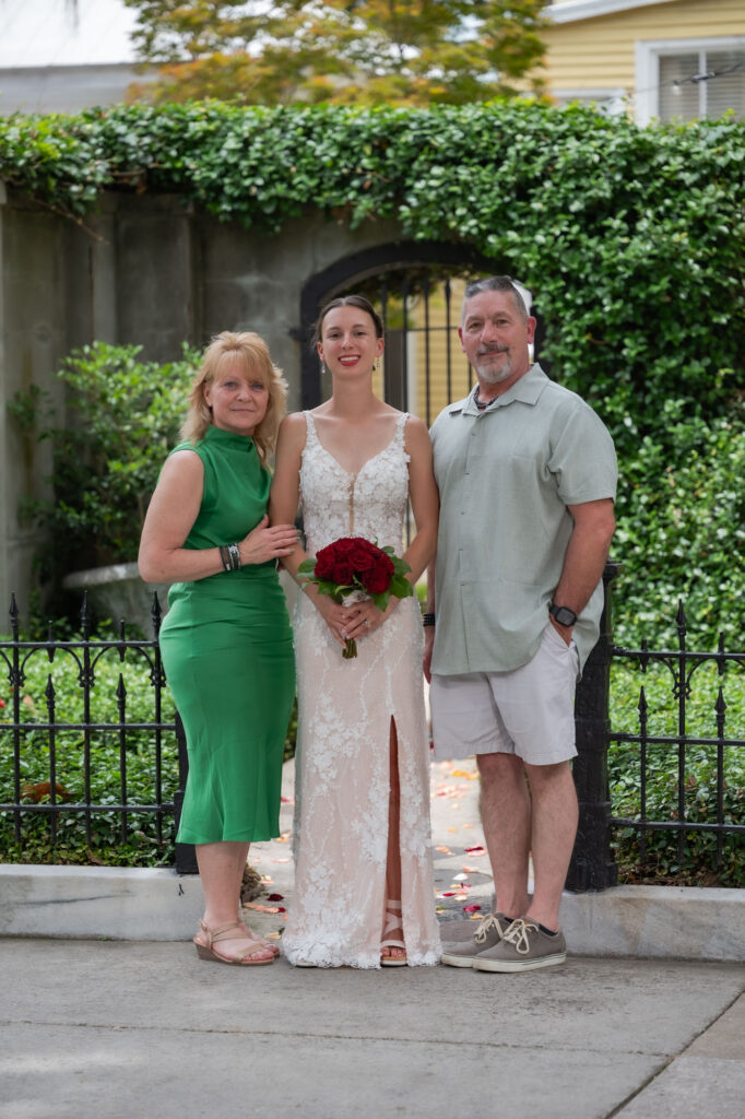 Three people posing outdoors: a bride in a white lace gown holding a red bouquet, flanked by a woman in green and a man in light gray shirt and shorts.