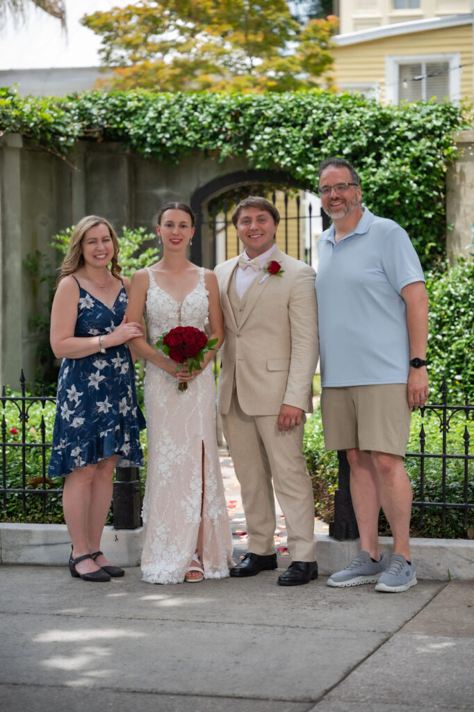 Bride in a white lace gown holds a red bouquet, standing with groom in a beige suit and two guests outdoors.