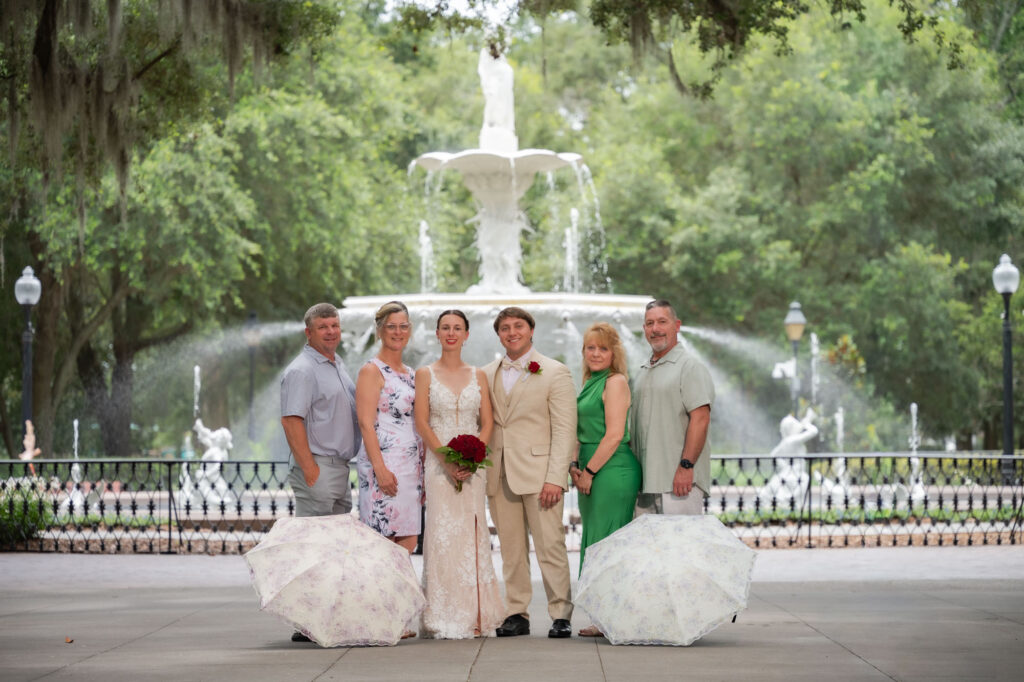 Bride in a lace wedding dress posing with family in front of a park fountain; two lace parasols lie on the ground nearby.