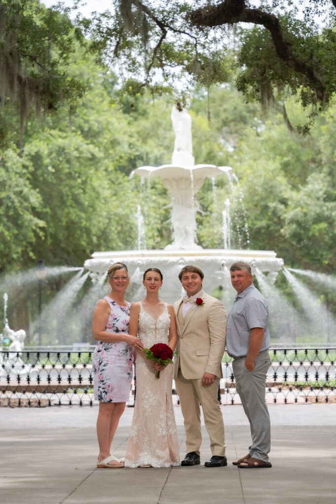 Bride in a lace wedding gown holding a red bouquet, flanked by two adults in front of a fountain in a park setting.