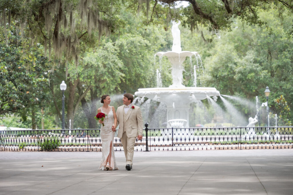 Bride in a white lace gown and groom in a tan suit walk hand in hand in a park with a large white fountain behind them.