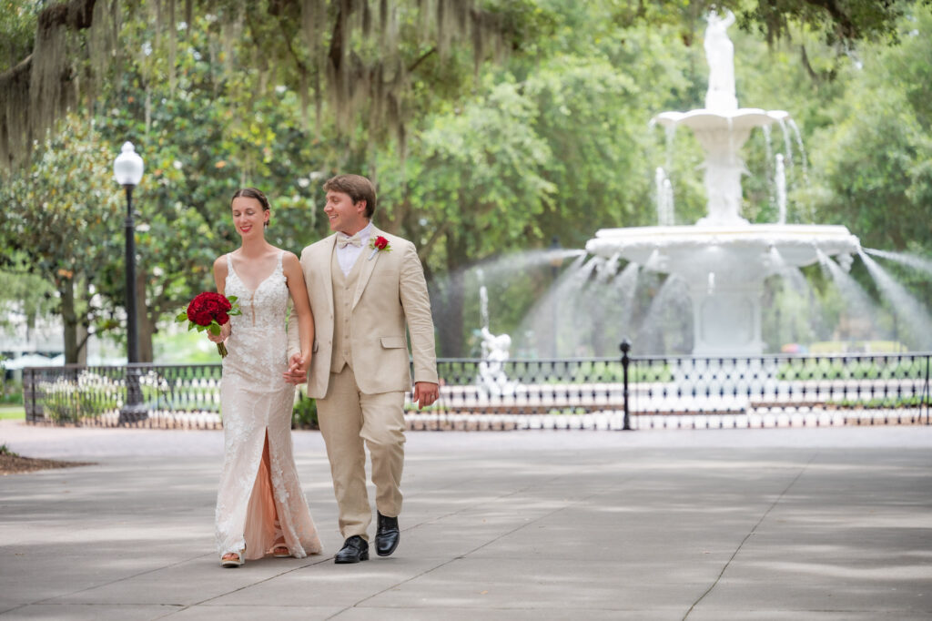 Bride in a white lace gown carries a red bouquet as she walks arm-in-arm with a groom in a tan suit, smiling, in a park with a decorative fountain in the background.