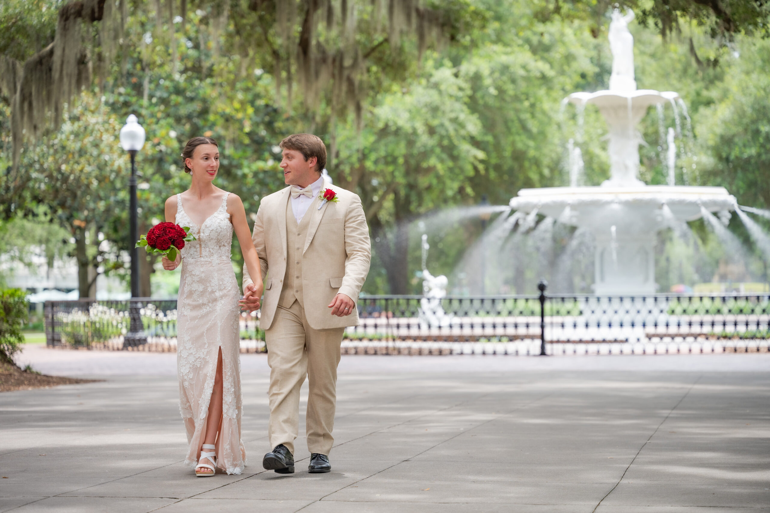 Bride in a lace wedding dress holding a red bouquet walks beside a groom in a tan suit along a park path with a fountain in the background.
