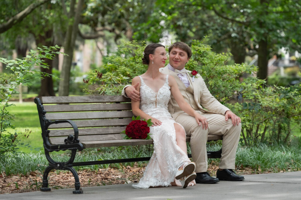Bride in a lace white wedding dress and groom in a beige suit sit close together on a park bench, red bouquet resting on her lap beneath greenery.