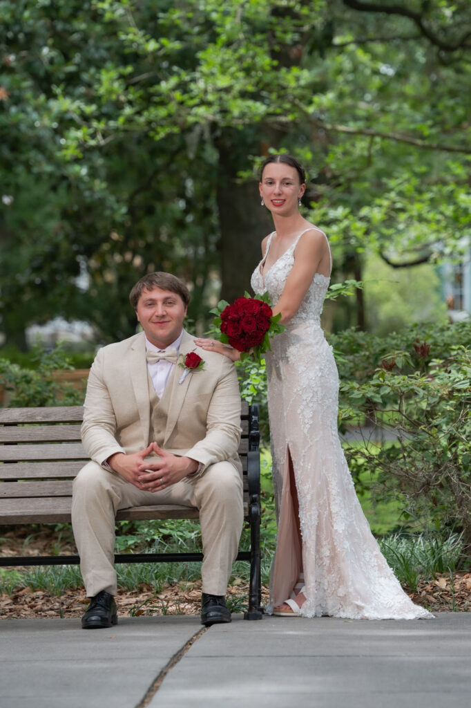 Bride in a lace wedding dress stands beside a seated groom in a beige suit on a park bench, holding a red rose bouquet.