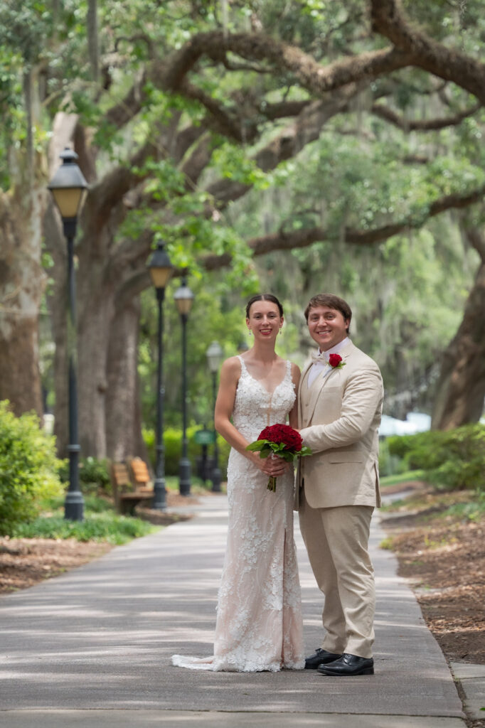 Bride in a lace wedding gown and a groom in a beige suit pose on a tree-lined park path, the bride holding a red bouquet. (Wedding portrait) | Outdoor park scene with couple smiling for photos under large trees and lamp posts behind them.