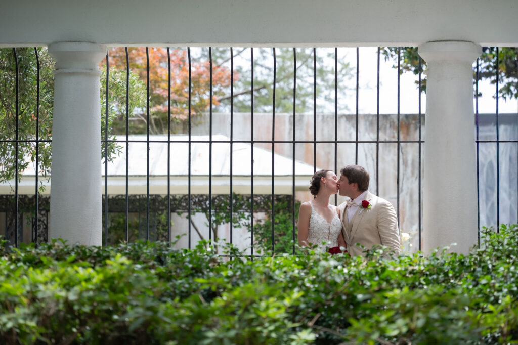 Bride and groom kiss behind a decorative iron fence with white columns and green shrubbery in the foreground.