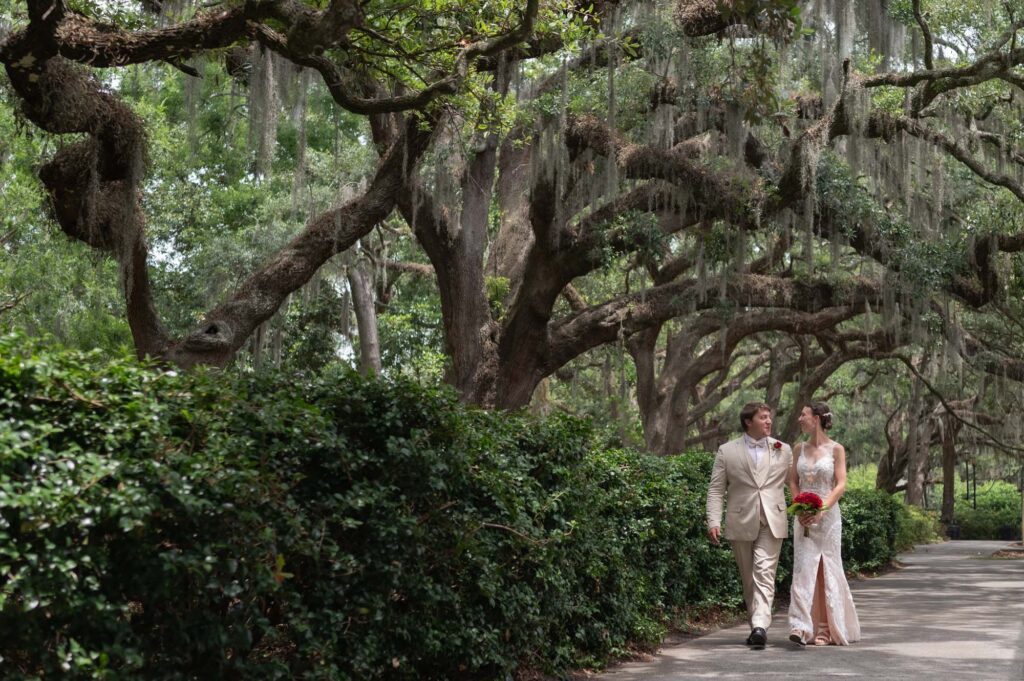 Bride and groom walking along a path in a lush garden with hanging Spanish moss and large trees, the bride holding a red bouquet and wearing a lace gown.