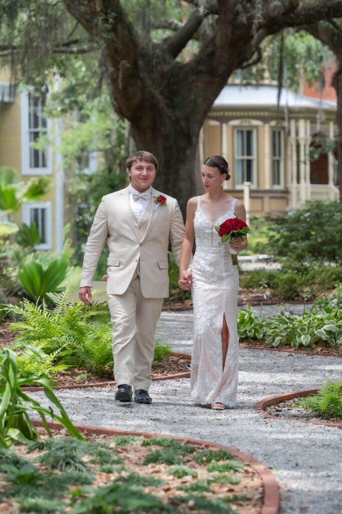 Bride in a white lace gown and groom in a light beige suit walk hand in hand along a garden path, holding a red bouquet with a large tree and a decorative house in the background.