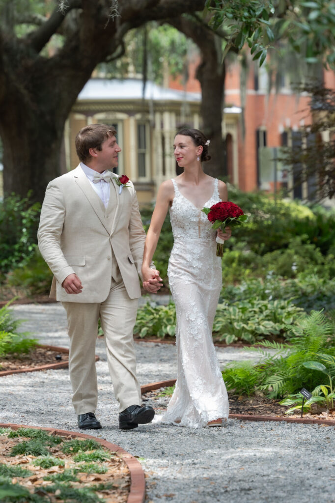 Bride in a lace wedding gown walks hand-in-hand with a groom in a beige suit along a garden path, holding a bouquet of red roses.