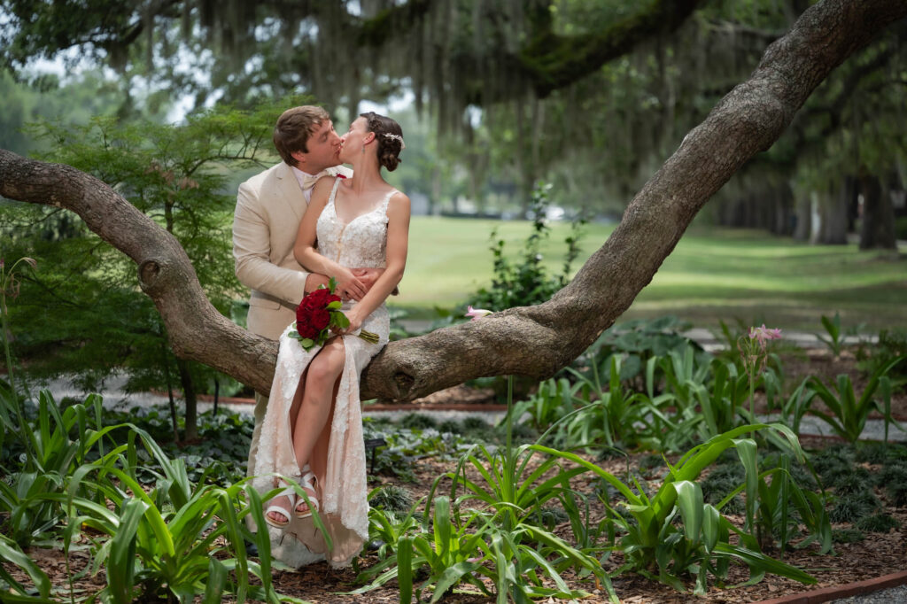 Bride and groom in wedding attire kiss while seated on a large tree branch in a garden, the bride holding a red bouquet.