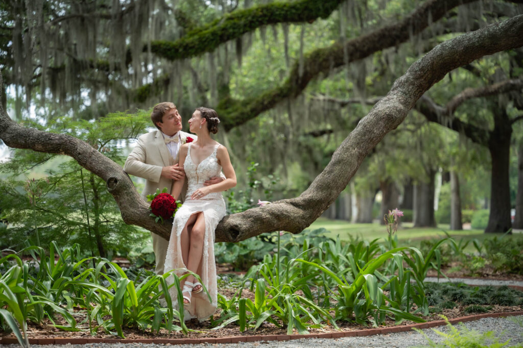 Bride in lace wedding dress and groom in a beige suit sit on a thick tree branch, holding a red bouquet nearby in a lush garden.