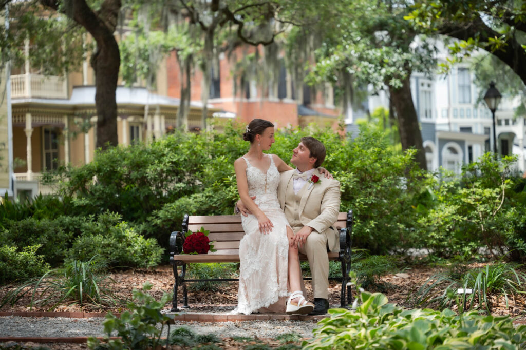 Bride in a lace wedding dress and groom in a beige suit sit closely on a park bench, with a red rose bouquet on the bench amid lush garden greenery.