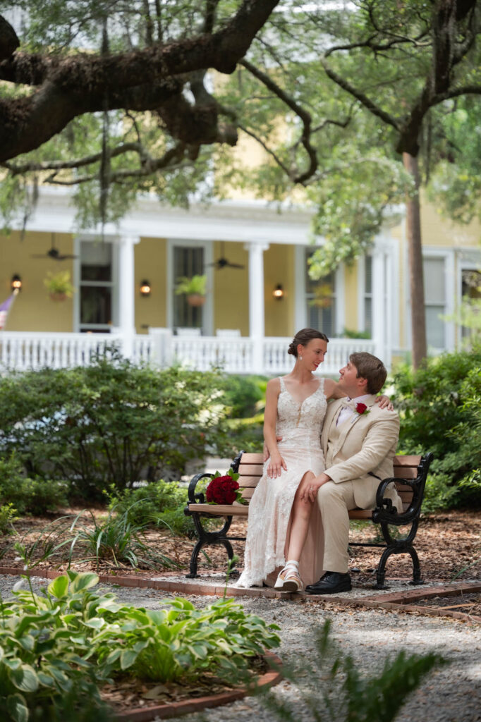 Bride in a lace white dress and groom in a beige suit sit close together on a park bench in a lush garden with a yellow house backdrop.