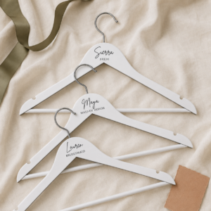 Three white hangers labeled for a wedding party on a beige fabric: Sierra (Bride), Maya (Maid of Honor), and Lauren (Bridesmaid).